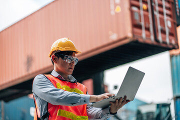 Asian technician dock worker in protective safety jumpsuit uniform and with hardhat and use laptop computer at cargo container shipping warehouse. transportation import,export logistic industrial