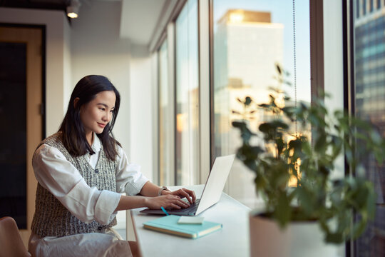 Young smiling Asian business woman employee or student working on laptop in corporate office. Happy professional businesswoman marketing manager using computer technology sitting at Generative AI