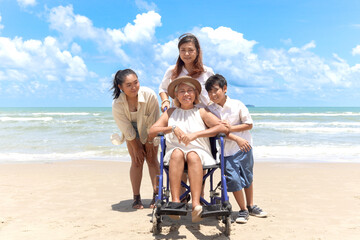 Happy disabled senior elderly woman in wheelchair spending time together with her family on tropical beach. Asian grandma, daughter and grandchild boy resting and relaxing on summer holiday vacation.