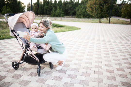 Young mother taking a selfie with her infant. The baby sits in a stroller. Mom is on maternity leave. Stylish mom in a knitted sweater and white trousers, baby in a pink eco knitted combination