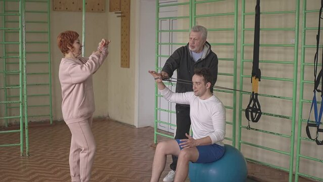 caucasian man is doing fitness using TRX loops in the gym, personal trainer is watching a sporty young man. a woman takes pictures on a smartphone. High quality Full HD video recording Generative AI