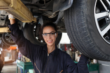 Beautiful female auto mechanic checking wheel tires in garage, car service technician woman repairing customer car at automobile service, inspecting vehicle underbody and suspension engine system.