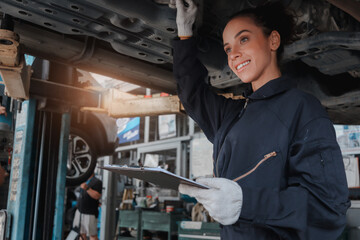 Beautiful female auto mechanic checking wheel tires in garage, car service technician woman repairing customer car at automobile service, inspecting vehicle underbody and suspension engine system.