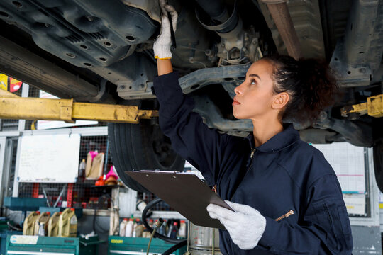 Beautiful female auto mechanic checking wheel tires in garage, car service technician woman repairing customer car at automobile service, inspecting vehicle underbody and suspension engine system.