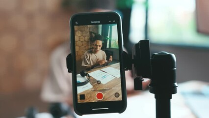 Young charismatic African American man blogger is recording advertisement on mobile phone camera mounted on tripod and talking about benefits of new smartphone sits at desk in home office.