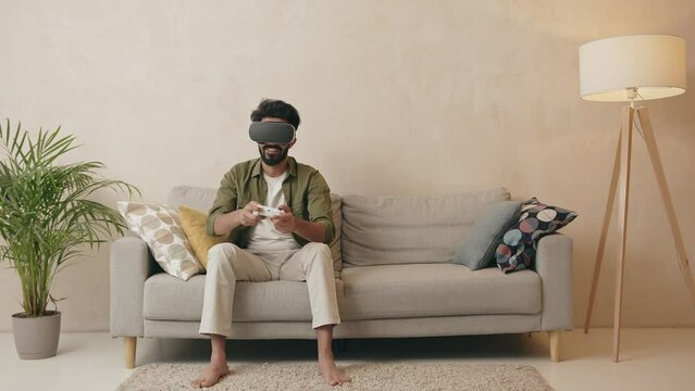 Excite indian man playing online game with vr glasses and controllers on sofa Generative AI