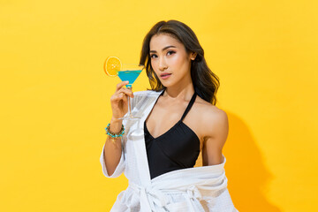 Sexy Asian woman wearing summer swimsuit and spa bathrobe holding cocktail drink in yellow color isolated background studio shot