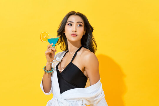 Sexy Asian woman wearing summer swimsuit and spa bathrobe holding cocktail drink in yellow color isolated background studio shot