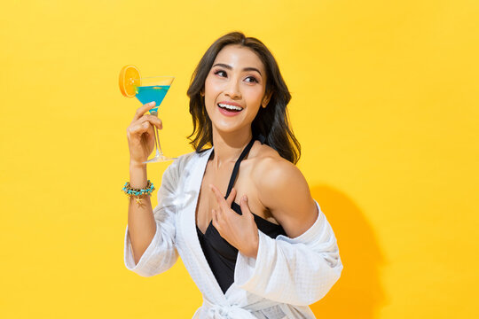 Smiling pretty Asian woman wearing summer swimsuit and spa bathrobe holding cocktail drink in yellow color isolated background studio shot