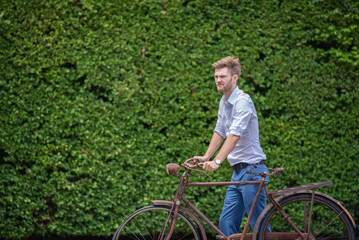 Man in white shirt blue jeans stand and pose with Old decay bicycle on green vine climbing garden wall outdoor. Rust Classic bike old bicycle on green garden wall retro style.