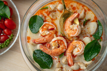 Tom Yam Kung or Tom Yum soup is Thai hot spicy soup shrimp or prawn with lemon grass, lemon, galangal, mushrooms, coconut milk and chili in bowl on wooden table top view, famous Thai food cuisine.
