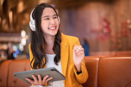 successful attractive smart business casual Asian female woman using tablet device working online remotely in cafe mall while listening music from headphone carefree working on holiday weekend