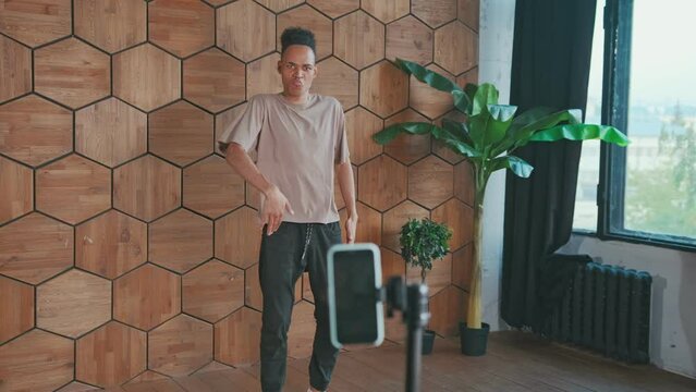 Young energetic African American man performs fashionable dance recording video on mobile phone camera mounted on tripod dressed ina casual style located in room near wooden wall. Blogging, challenge