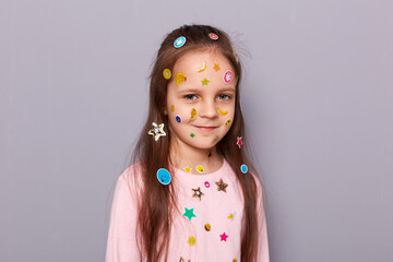 Positive cute little girl covered colorful kid's stickers standing isolated over gray background looking at camera with calm expression and charming smile.