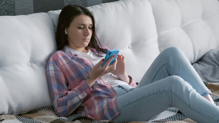 Millennial caucasian girl checking social media holding smartphone at home. Young woman using mobile phone app playing game, shopping online, ordering delivery relax on sofa.