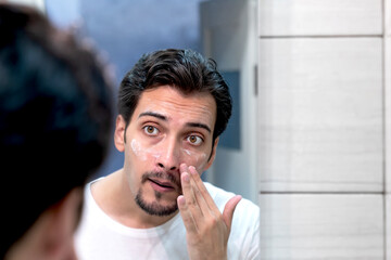 Handsome man looking at mirror and applying moisturizing cream on cheeks in bathroom. Groomed young guy doing skincare morning routine after taking a shower