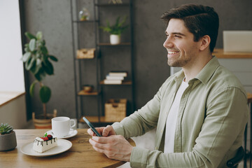 Side viw young minded happy smiling man wears casual clothes sits alone at table in coffee shop...