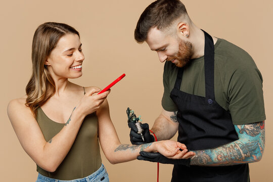 Tattooer master artist tattooed man wears green t-shirt apron hold machine black ink in jar equipment to make tattoo art body isolated on plain beige background. Woman take photo on mobile cell phone.