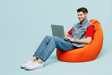 Full body young IT man he wear denim vest red t-shirt casual clothes sit in bag chair hold use work on laptop pc computer isolated on plain pastel light blue cyan background studio. Lifestyle concept.