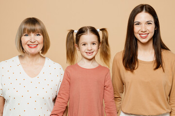 Close up happy smiling women wear casual clothes with child kid girl 6-7 years old. Granny mother daughter holding hands looking camera isolated on plain beige background. Family parent day concept.