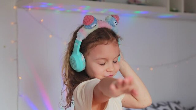 Cute 7 year old girl with headphones listening to music and dance in her room against a background of colored light, portrait. The concept of free time and hobbies of a modern child
