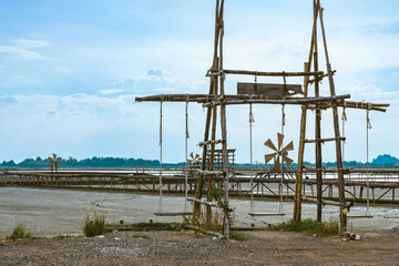 Fototapeta na wymiar Wooden swings for relaxing and taking photos and wind wheel at the salt pan view with blue sky background in summer time of Thailand. Beautiful landscape of salt field.Traditional salt farming culture