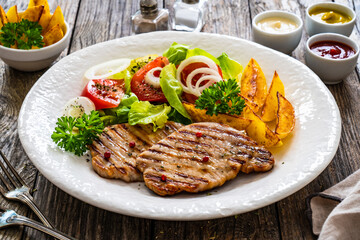 Barbecued pork chops with fried potatoes and fresh vegetables on wooden table
