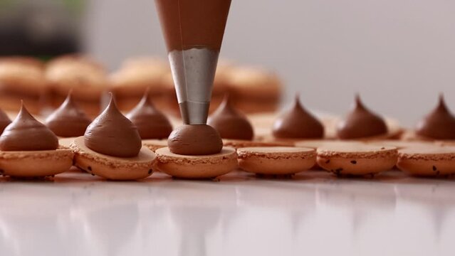 Making french desert macarons. Chef hands with confectionery bag add cream to the ready halves of brown chocolate macaroons at pastry shop. cooking, food and baking concept