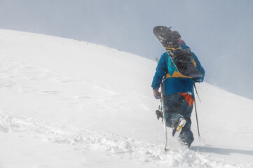 man with ski equipment and a snowboard climbs a snowy mountain. Blue sky and snowy mountain in the...