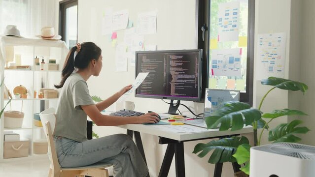Side view of professional young Asia girl IT development programmer typing on keyboard coding programming fixing data code on computer screen and laptop on table in workroom at house office.
