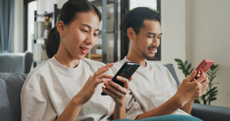 Young Asian couple sit on couch spend time together have fun use smartphone devices online shopping at home. Happy Young husband and wife laugh relax with phone, Lifestyle domestic activity concept.