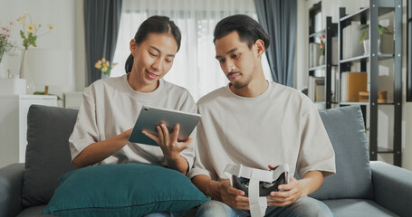 Young Asian couple sit on couch hold digital tablet and wear virtual reality glasses are watching and show imagine spend time together have fun at home on weekend. Lifestyle tech innovation concept.