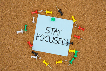 Stayed focus text on light blue post-it paper pinned on bulletin cork board surrounding by multi...