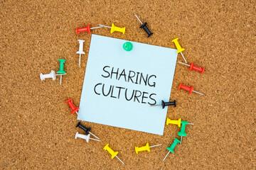Sharing cultures text on light blue post-it paper pinned on bulletin cork board surrounding by multi color pins. This message can be used in business concept about cultures.