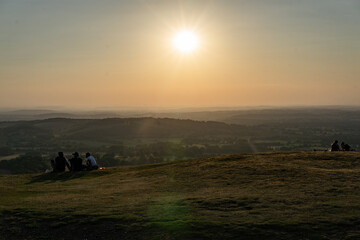Naklejka na ściany i meble A group of people admiring the beauty of nature at sunrise, watching as the sun rises above a distant horizon over a picturesque hill. View from British camp Malvern Hills