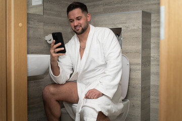 A man in a white coat with a smile on his face and a mobile phone in his hand sits in the toilet...