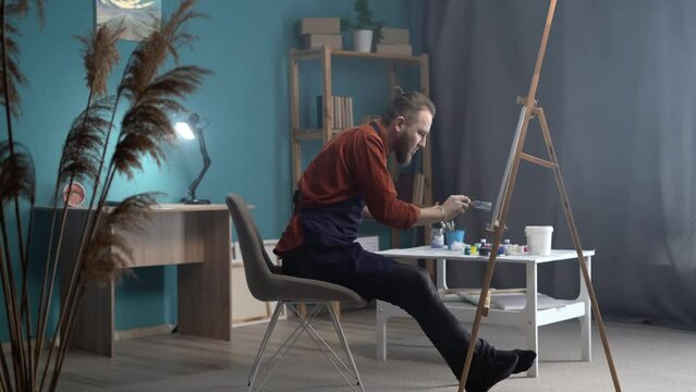 portrait of a bearded millennial artist working in an apron while sitting on an abstract painting, uses a brush to create a modern side view painting. canvas on an easel in a creative studio.