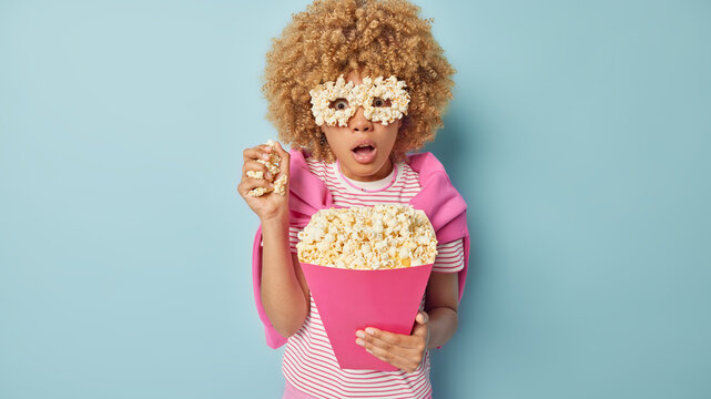 Photo of curly haired young woman watches horror film eats popcorn feels impressed makes eyeglasses from food dressed in t shirt and jumper tied over shoulders isolated over blue Generative AI