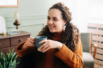Cozy Winter Mornings: Happy Woman Enjoying Warm Drink and Daydreaming at Home. Warmth and Relaxation: Young Woman Holding a Cup of Coffee and Daydreaming at Home.