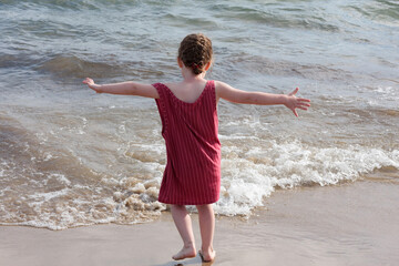 Fototapeta na wymiar little girl on the beach in a red dress with arms outstretched