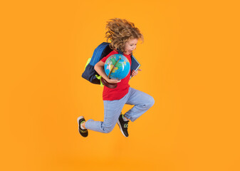 Fototapeta na wymiar Back to school. Full body of energetic primary school kid boy wearing casual clothes holding backpackand copybook globe and jumping against blue background.