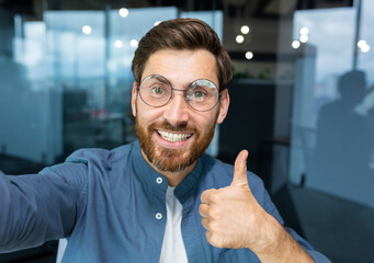 Obraz premium Close-up photo. A young freelancer man, a blogger in glasses, talks to the camera, smiles, shows a super hand, records a video, blogs.