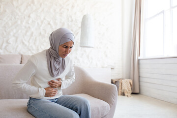 Unhappy young Arab woman suffering from stomach pain. She sits on the sofa and holds her stomach with her hands, bent over, grimacing. Needs medical help.