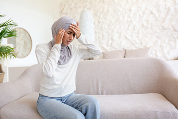 A young woman in a hijab is sitting on the sofa at home and holding her head with her hands,...