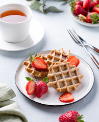 Homemade soft waffles with fresh strawberries and powdered sugar in a plate on a light background with a cup of tea. Traditional Belgian waffles. Healthy breakfast