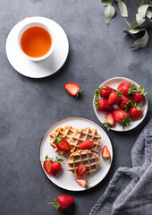 Homemade soft waffles with fresh strawberries and powdered sugar in a plate on a dark background with a cup of tea. Traditional Belgian waffles. Healthy vegetarian breakfast.