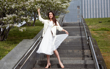 girl in a white dress stands on the stairs in a big city next to flowering trees in spring