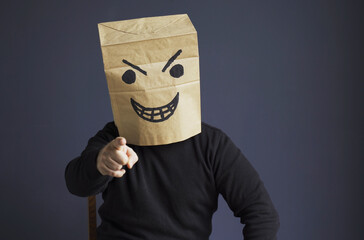 A man in a black turtleneck with a paper bag with an angry emoticon on his head shows his hand pointing forward.
