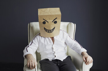 A man with a paper bag on his head, with a drawn angry smiley face, sits in a white chair.