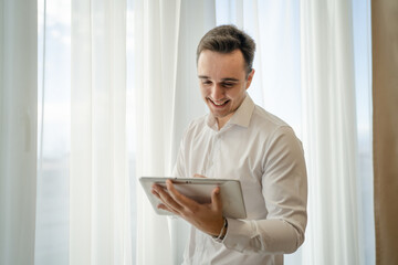 man wear white shirt hold cup of coffee and digital tablet at window